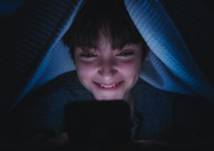 Happy teen girl using smartphone, hiding under blanket at nigh, social networks cocnept