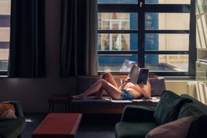 young woman in summer clothes lying on couch reading a book in a warm daylight under a big window